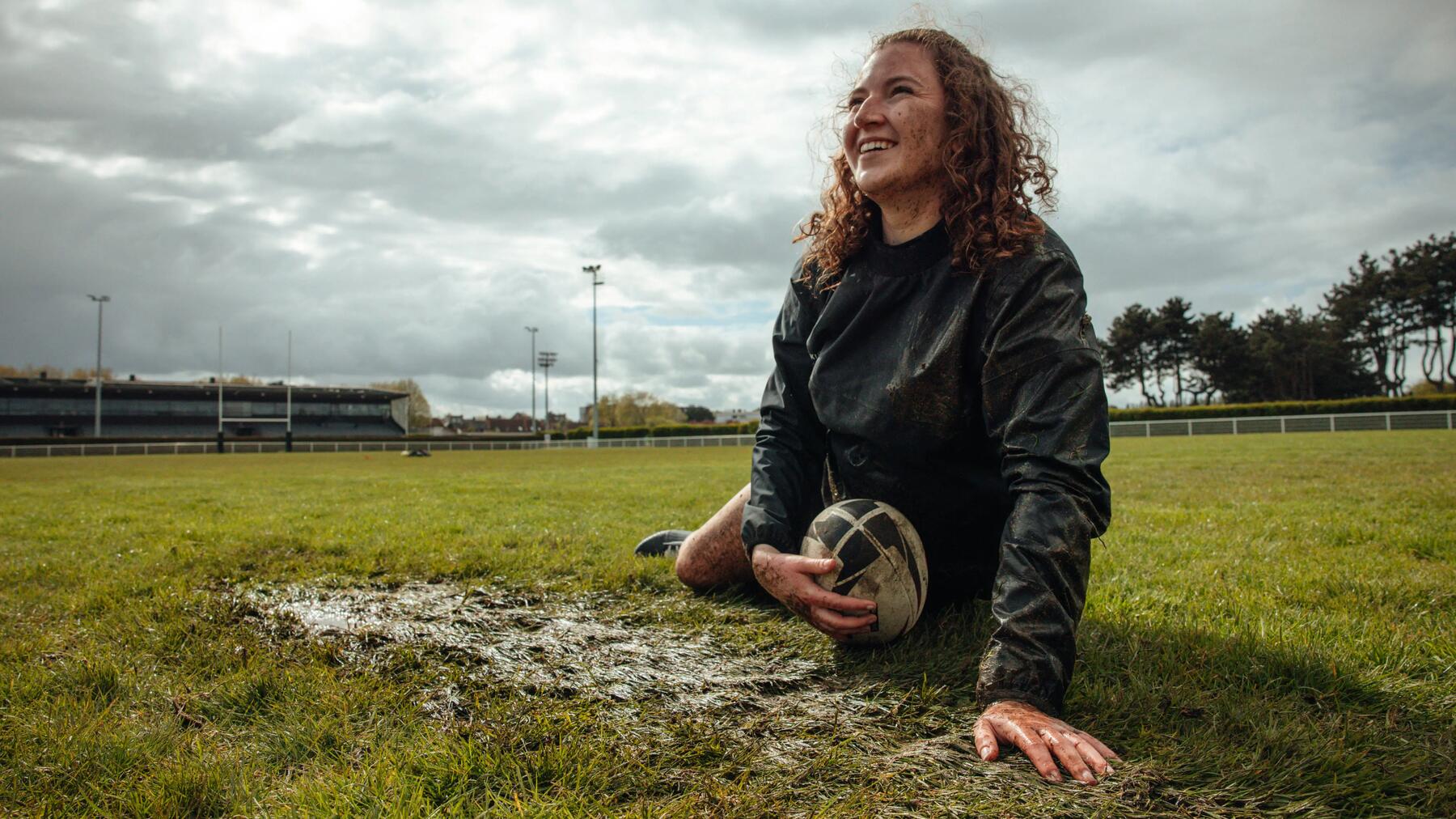 Frau sitzt auf matschigem Rugbyfeld, lächelnd, in dunkler Kleidung, bedeckt mit Schmutz, bewölkter Himmel, grüne Wiese.