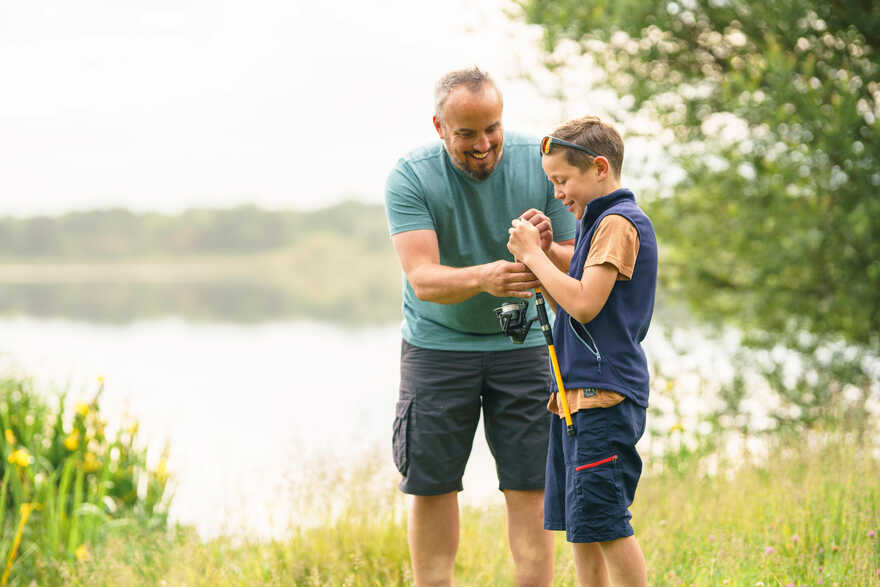 Oče pomaga sinu pri pripravi ribiške palice ob obali mirnega jezera, obdane z zelenjem.