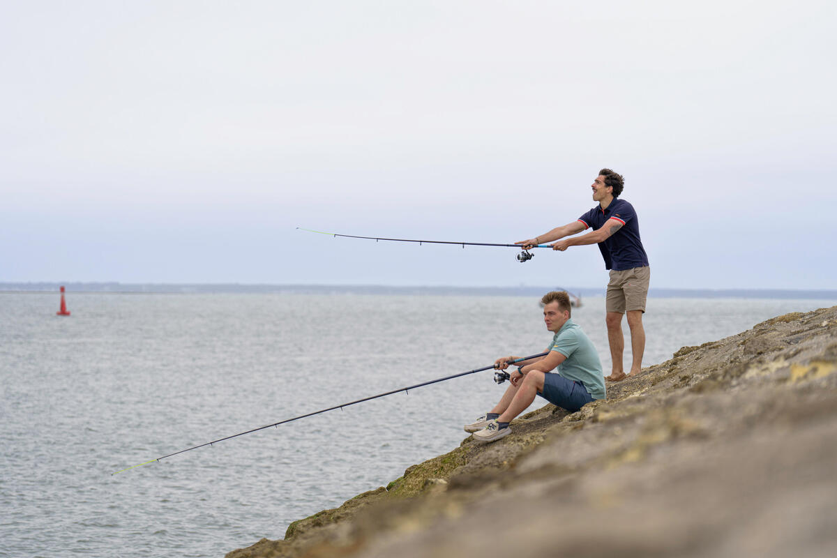 Dva mlada ribiča ribarita s kamnite obale ob morju s postavljenimi ribiškimi palicami med mirnim dnevom.