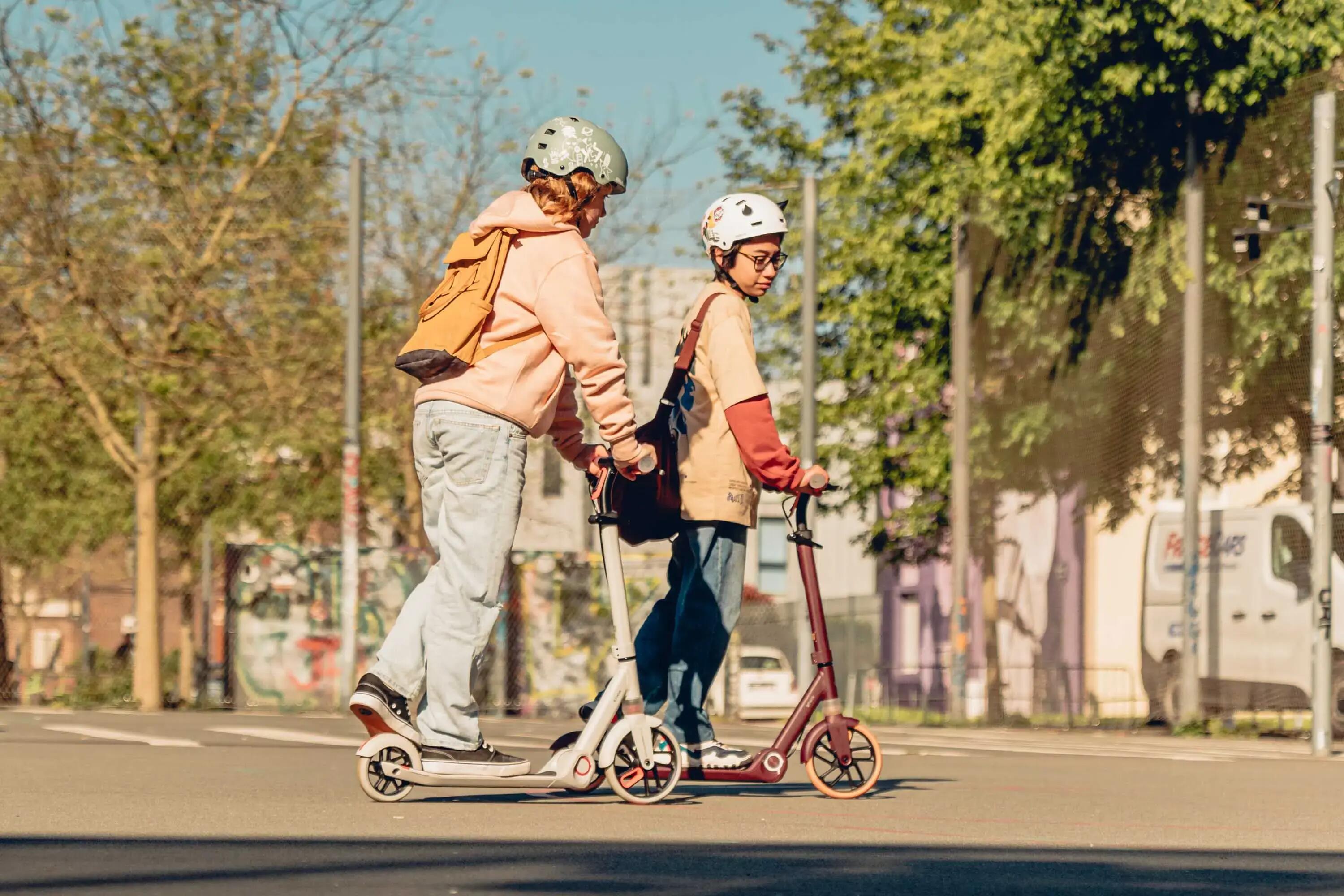 Zwei Jugendliche fahren mit Helmen auf E-Scootern auf einer asphaltierten Straße in der Stadt. Sie sind umgeben von Bäumen, Strommasten und Gebäuden.