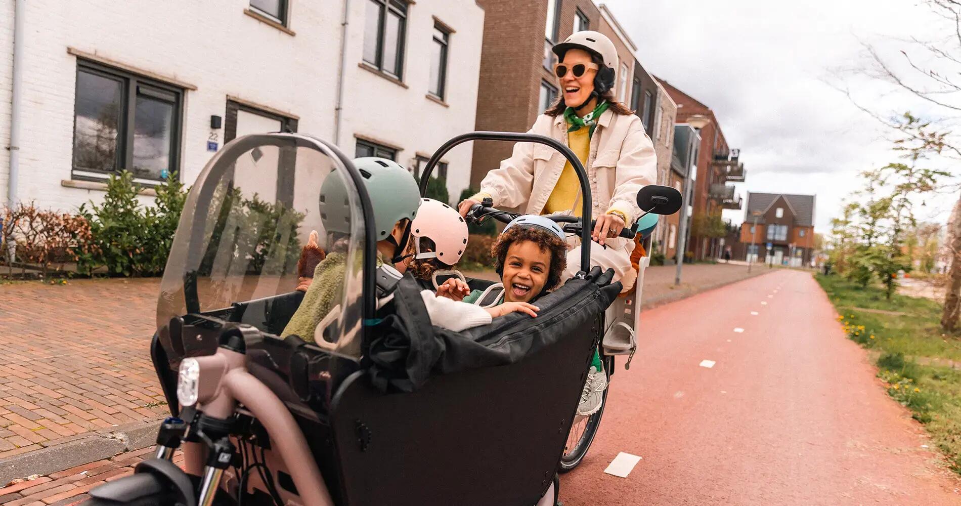 Eine Frau fährt ein Lastenfahrrad auf einem rötlichen Fahrradweg. In der vorderen Transportbox sitzen drei Kinder mit Fahrradhelmen, die fröhlich in die Kamera blicken oder miteinander interagieren. Im Hintergrund sind Wohnhäuser zu sehen.