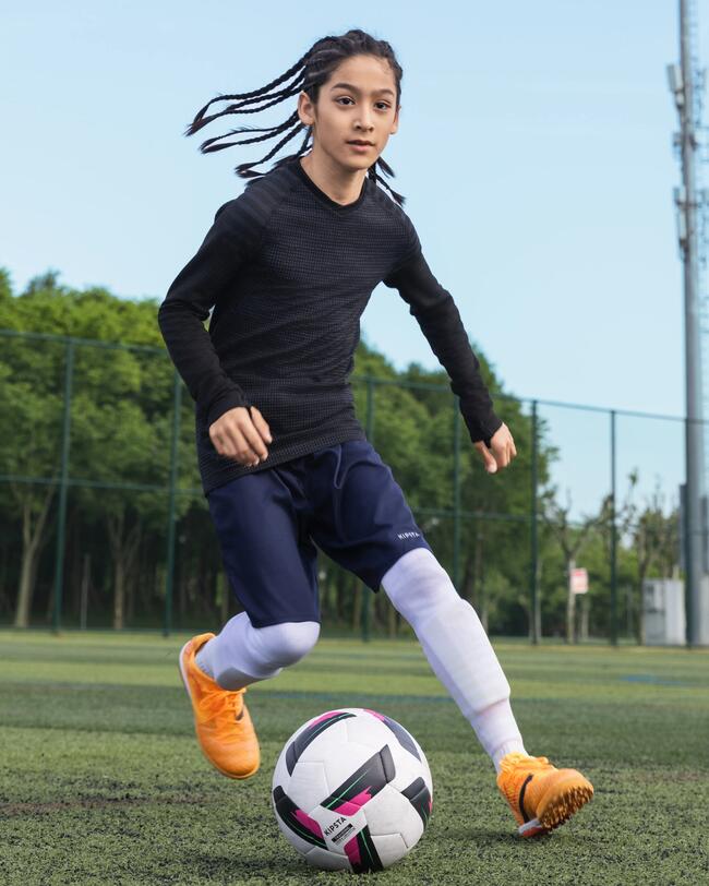 Junge im schwarzen langärmligen Keepdry Shirt dribbelt mit Fußball auf einem Fußballplatz und trägt dazu dunkelblaue Shorts, weiße Stutzen und orangene Schuhe. Bäume und  blauer Himmel im Hintergrund.