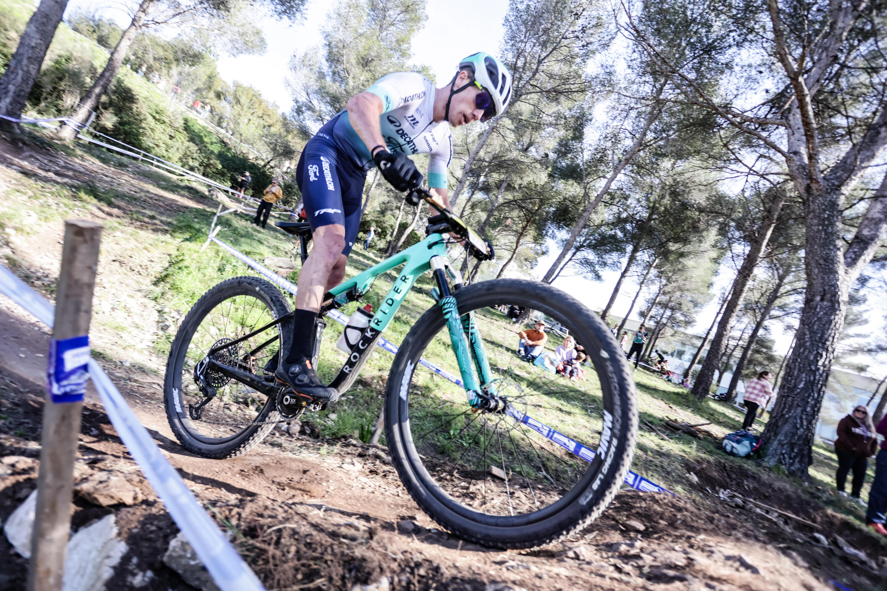 Ein Mountainbiker in Decathlon Rockrider Team-Bekleidung fährt ein türkisfarbenes Rockrider Mountainbike über einen erdigen Trail im Wald. Er befindet sich in einer Kurve, trägt einen Helm und eine Sportbrille, während im Hintergrund Bäume und Zuschauer zu sehen sind.
