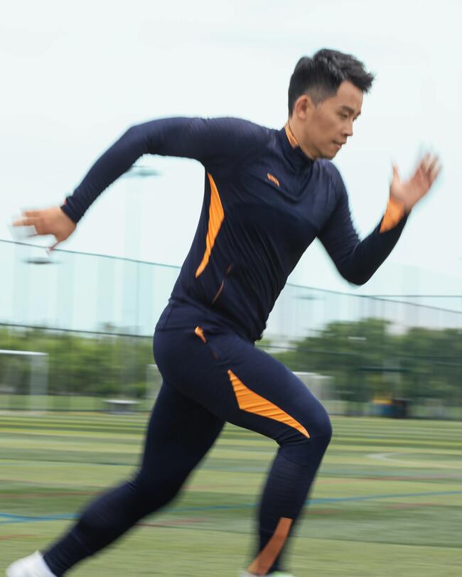 Sportler im CLR Sweatshirt in Dunkelblau mit orangefarbenen Streifen sprintet über grünes Spielfeld, glatte Struktur.