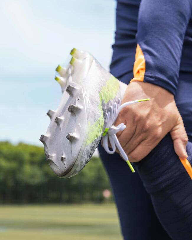 Fußballstollenschuh, silber und neongelb, auf grünem Feld. Person in dunkelblauer Sportkleidung dehnt Bein.