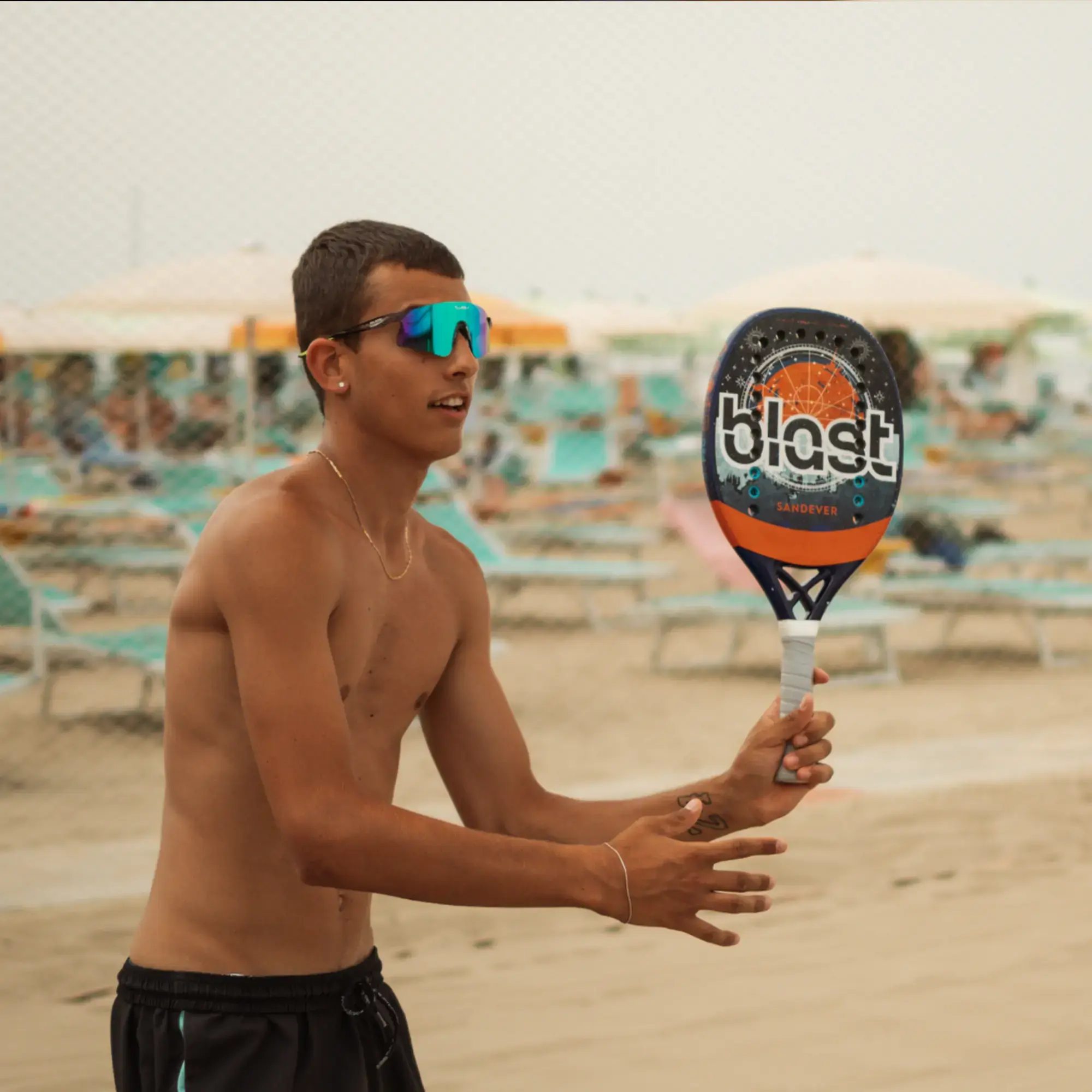 Beach-Tennis-Spieler mit spiegelnder Sport-Sonnenbrille hält einen Sandever Blast Beach-Tennis-Schläger spielbereit am Strand.