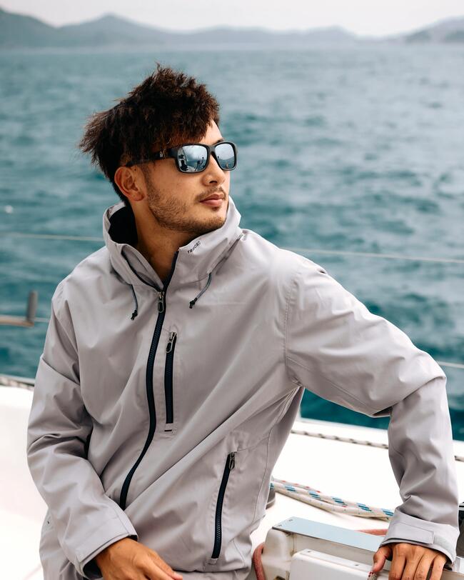 Mann mit grauer Jacke und Sonnenbrille auf einem Boot vor blauem Meer.