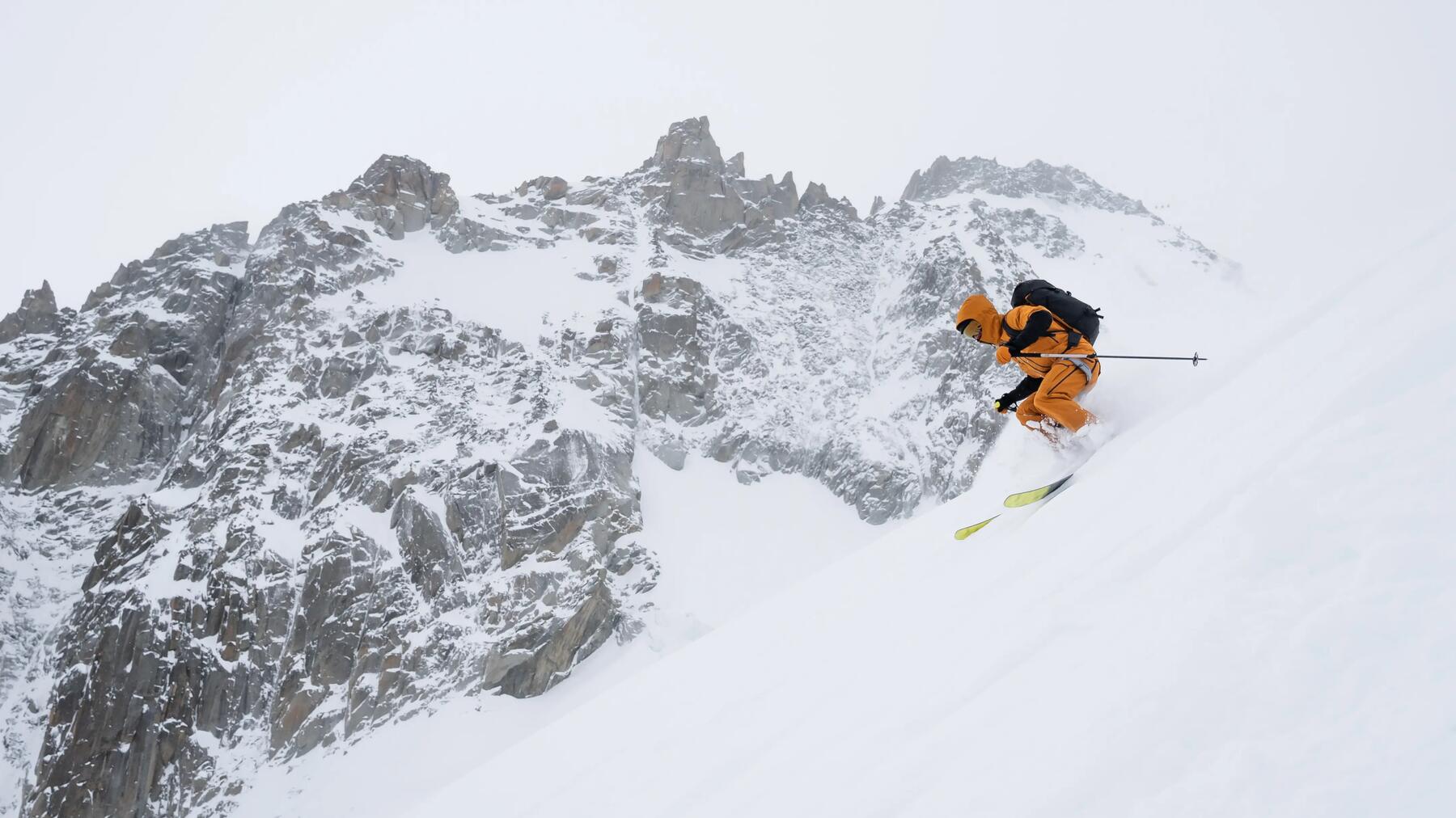 Skifahrer in gelber Skibekleidung und mit gelben Skiern beim Freeskiing auf der Piste mit Berg im Hintergrund.
