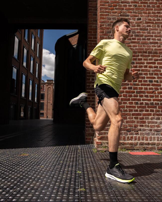 Läufer in gelbem Shirt, schwarzen Shorts auf gepflasterter Straße neben roten Backsteinmauern, blauer Himmel im Hintergrund.