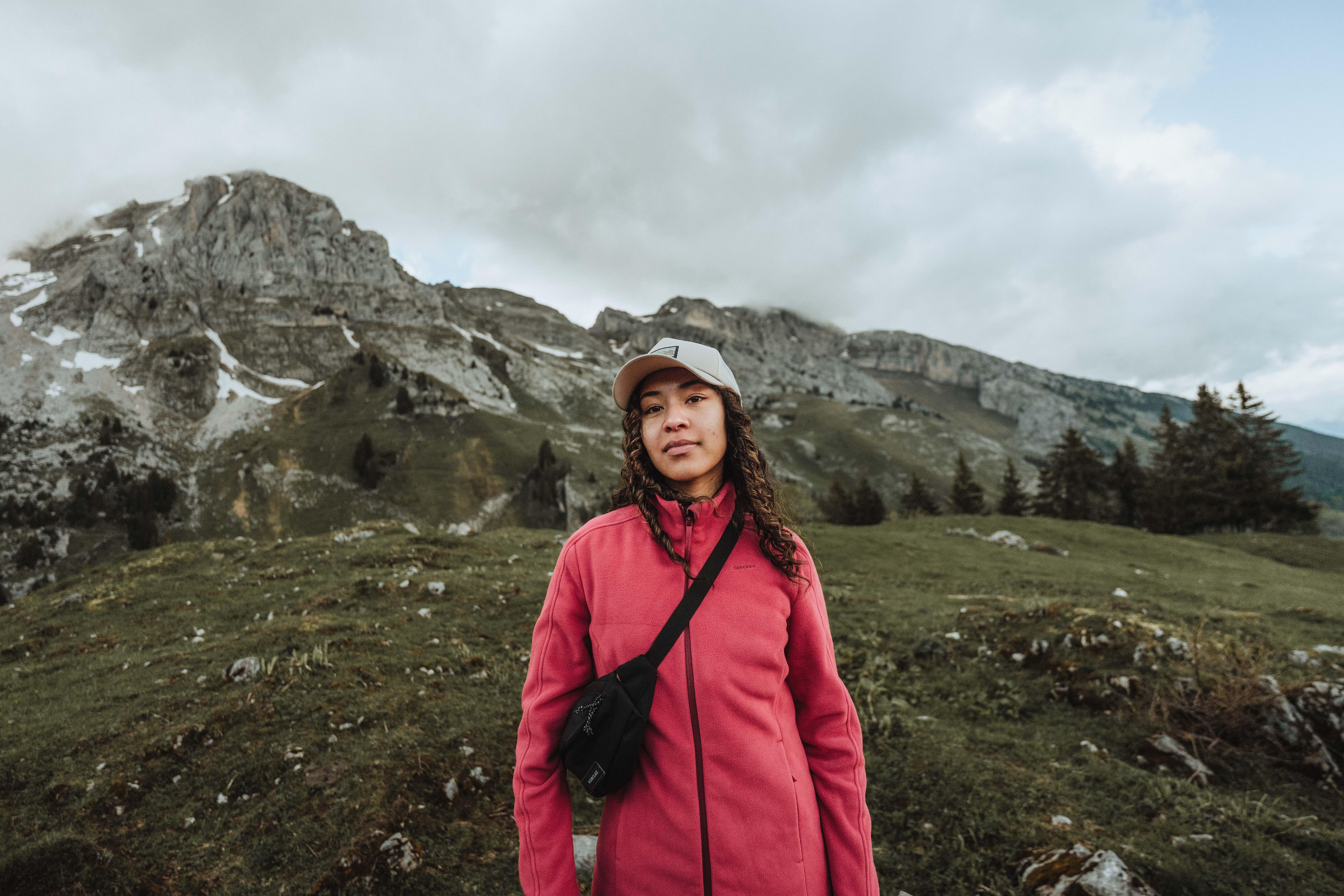 Woman wearing a pink hiking jacket and black crossbody bag in a mountainous landscape.