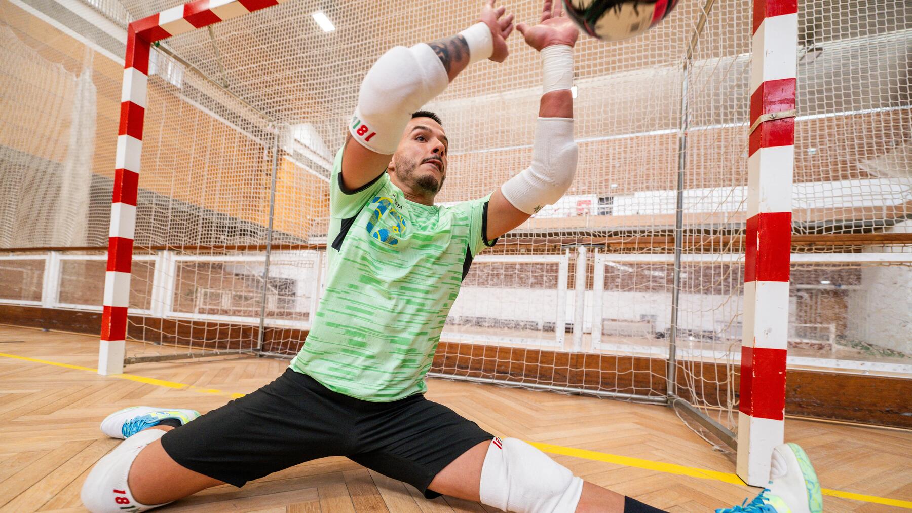 Ein Futsal-Torwart im hellgrünen Trikot macht vor dem Tor einen Spagat auf dem Hallenboden, um einen Ball abzuwehren.