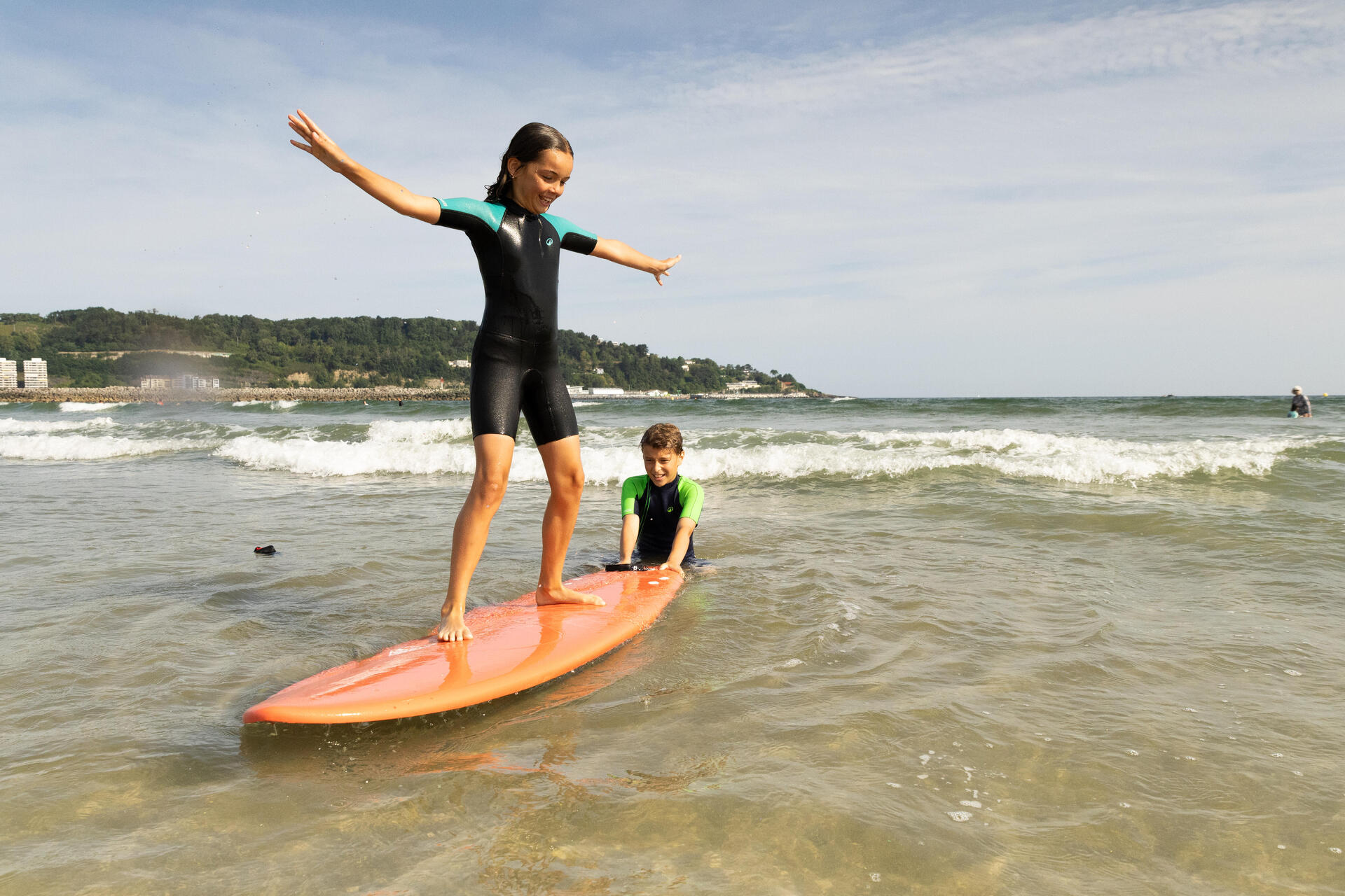 Dziewczynka w piance neoprenowej trenująca surfing na brzegu w towarzystwie chłopca pomagającego jej utrzymać się na desce