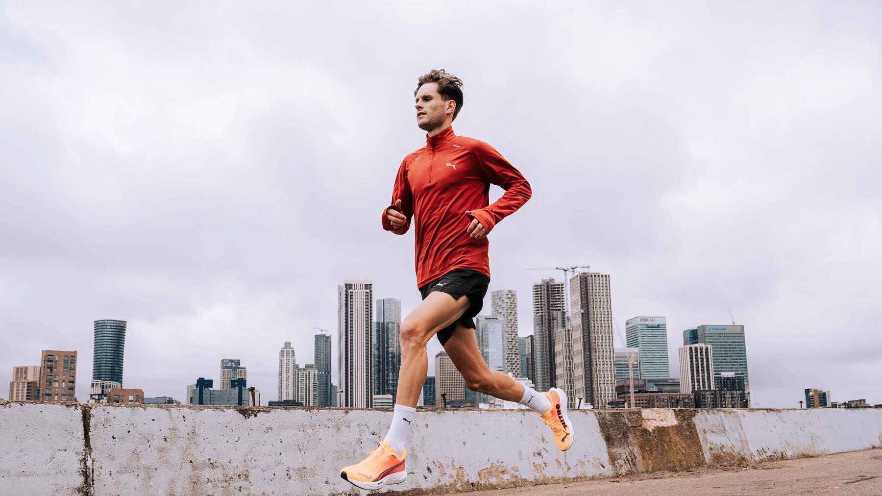 Mann in roter Jacke und schwarzen Shorts joggt auf Asphalt vor moderner Skyline, bewölkter Himmel, helle Schuhe, urbane Kulisse.