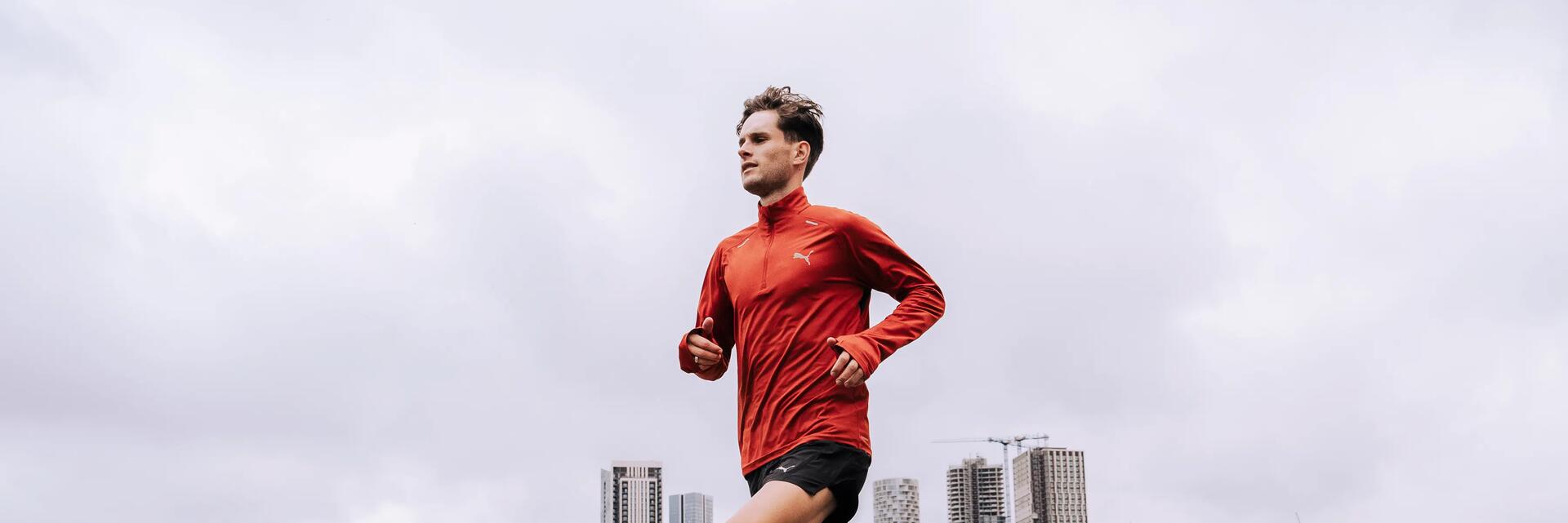 Mann im roten Sportshirt läuft vor grauen Hochhäusern, heller Himmel, orange Laufschuhe, raue Asphalttextur, moderne Skyline.