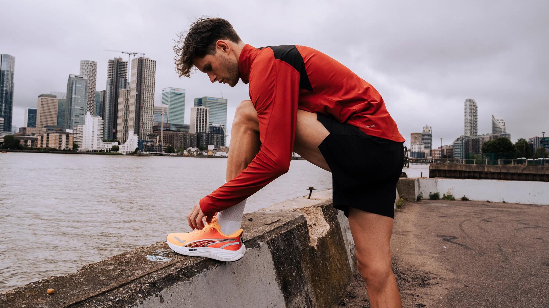 Mann in rotem Sportshirt bindet Turnschuh vor urbaner Skyline, graue Wolken, rauer Beton, orange-weißer Schuh.