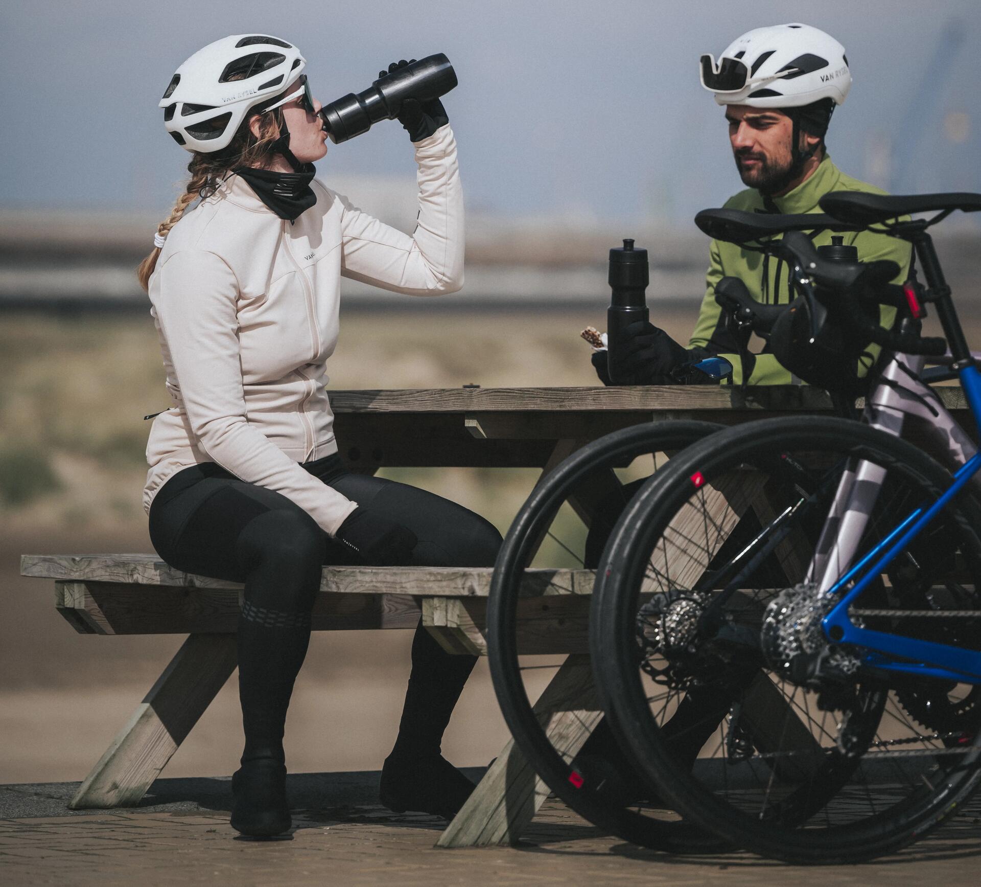 Two cyclists sat on a bench with their bikes, the lady is drinking from a water bottle