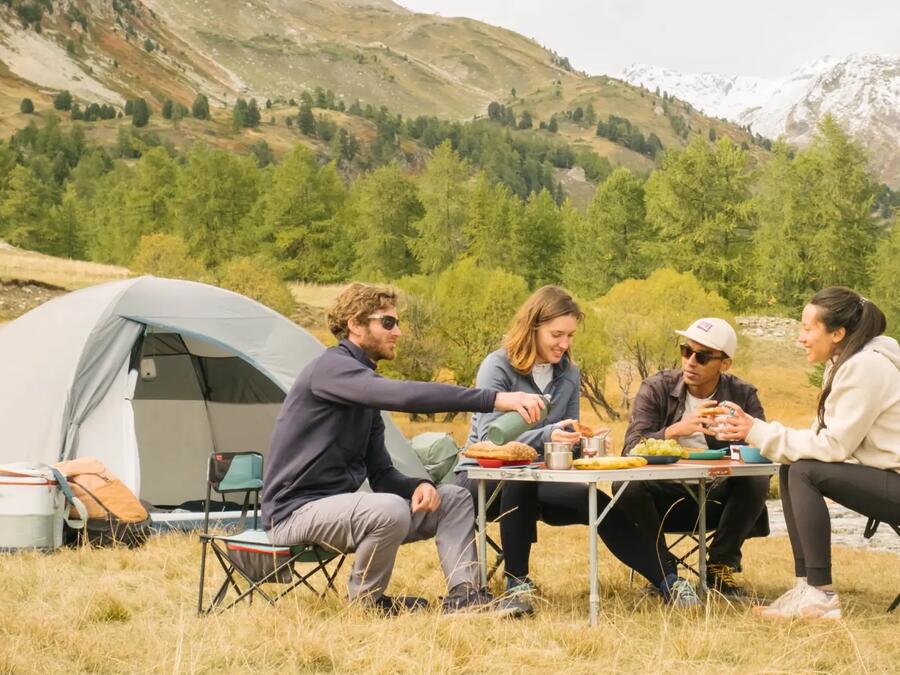 Four friends sat on camping chairs smiling and eating outside on a camping table