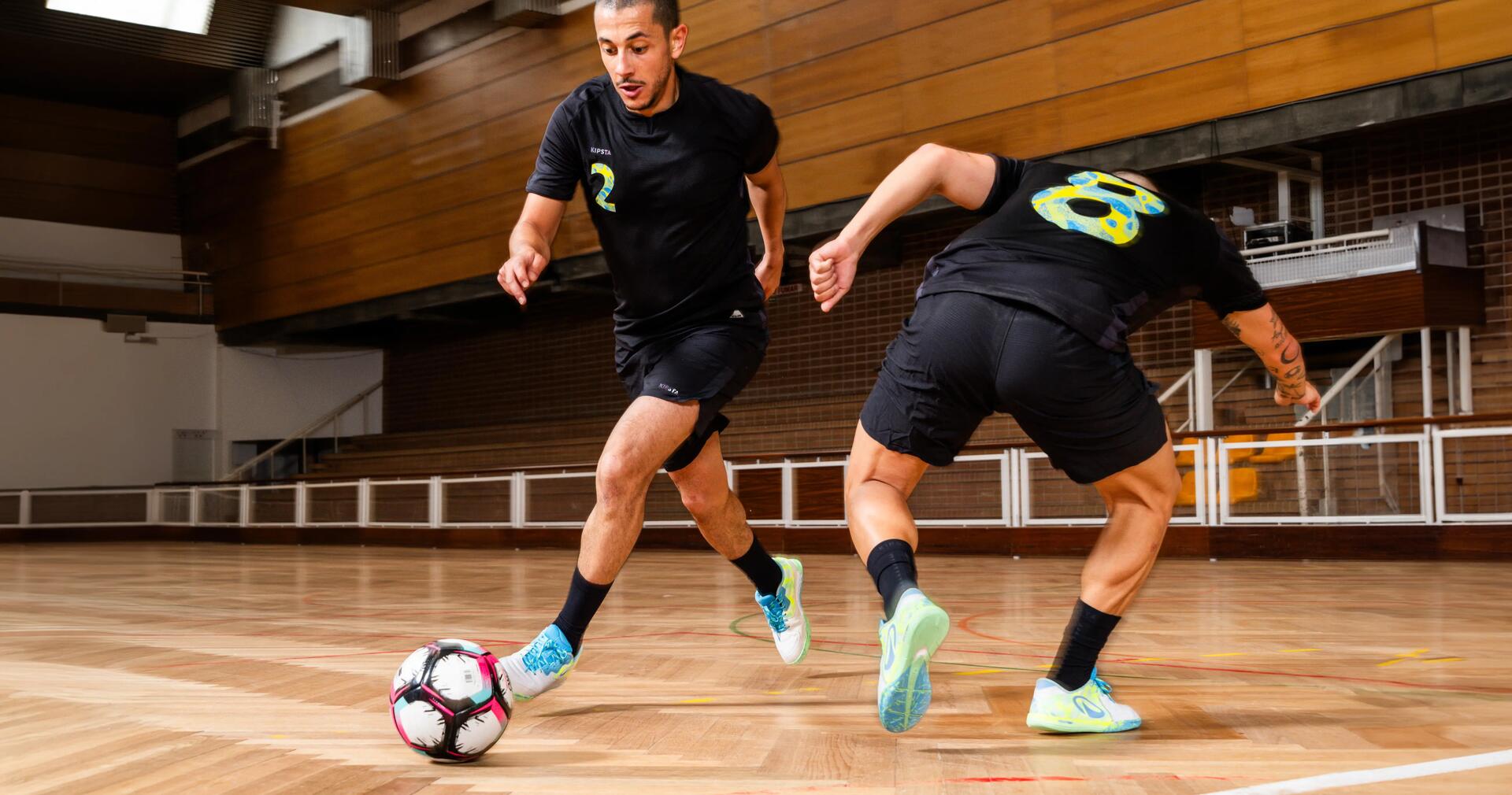 Zwei Männer spielen Futsal in einer Sporthalle, während einer von ihnen mit dem Ball am Fuß an seinem Gegenspieler vorbeidribbelt.