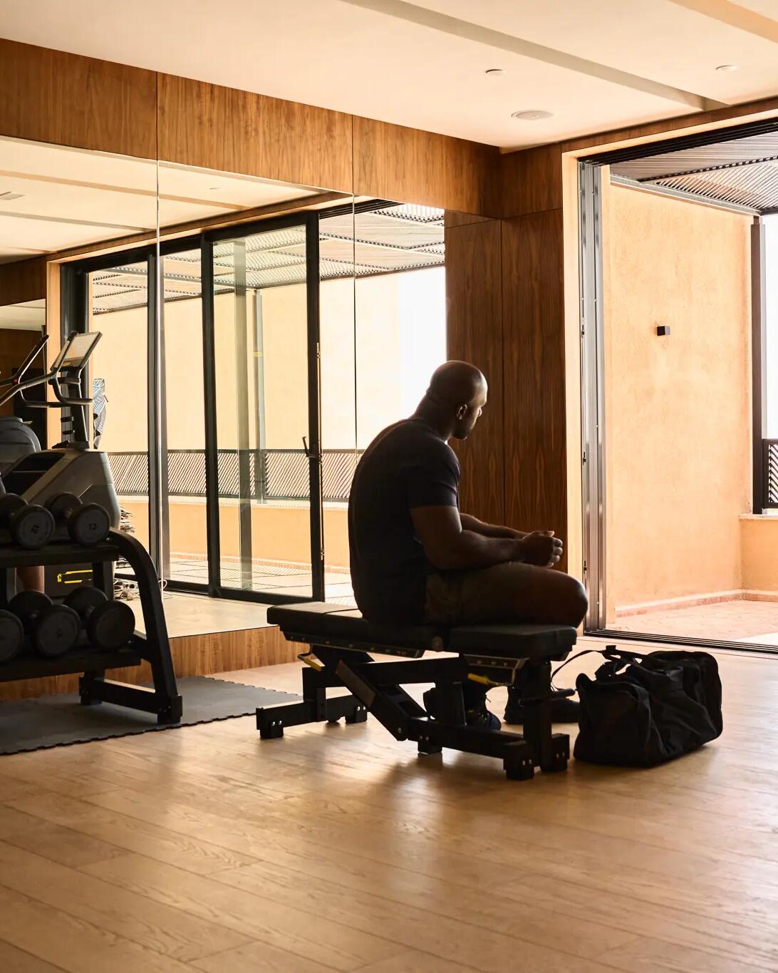 Ein Mann sitzt in Silhouette auf einer Trainingsbank in einem hellen Fitnessraum vor einer Fensterfront.