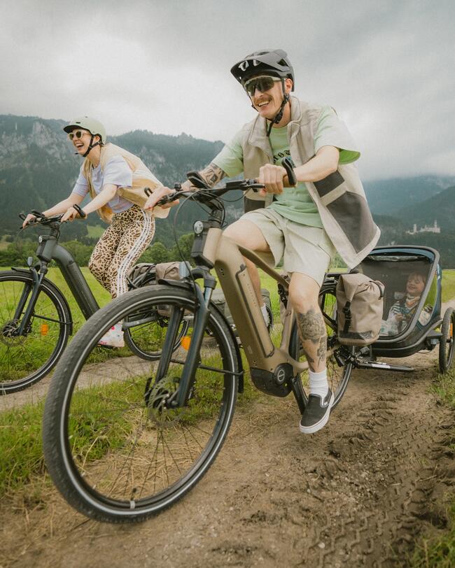 Familie fährt auf zwei E-Bikes über Feldweg. Am vorderen braunen Fahrrad ist ein Kinderanhänger befestigt.