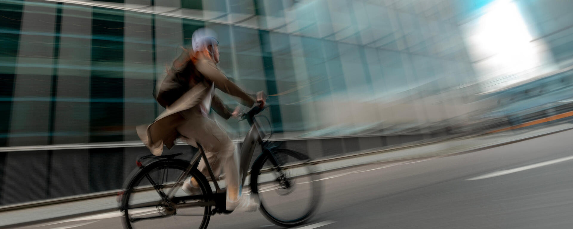Person mit Helm auf schwarzem Fahrrad, verschwommene Bewegung, moderne Glasgebäude im Hintergrund, graue Straße.