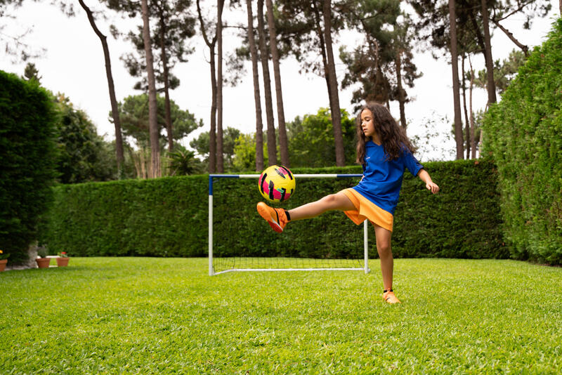 Dečak u narandžasto-plavom fudbalskom kompletu stoji na fudbalskom terenu i sprema se da šutne loptu.