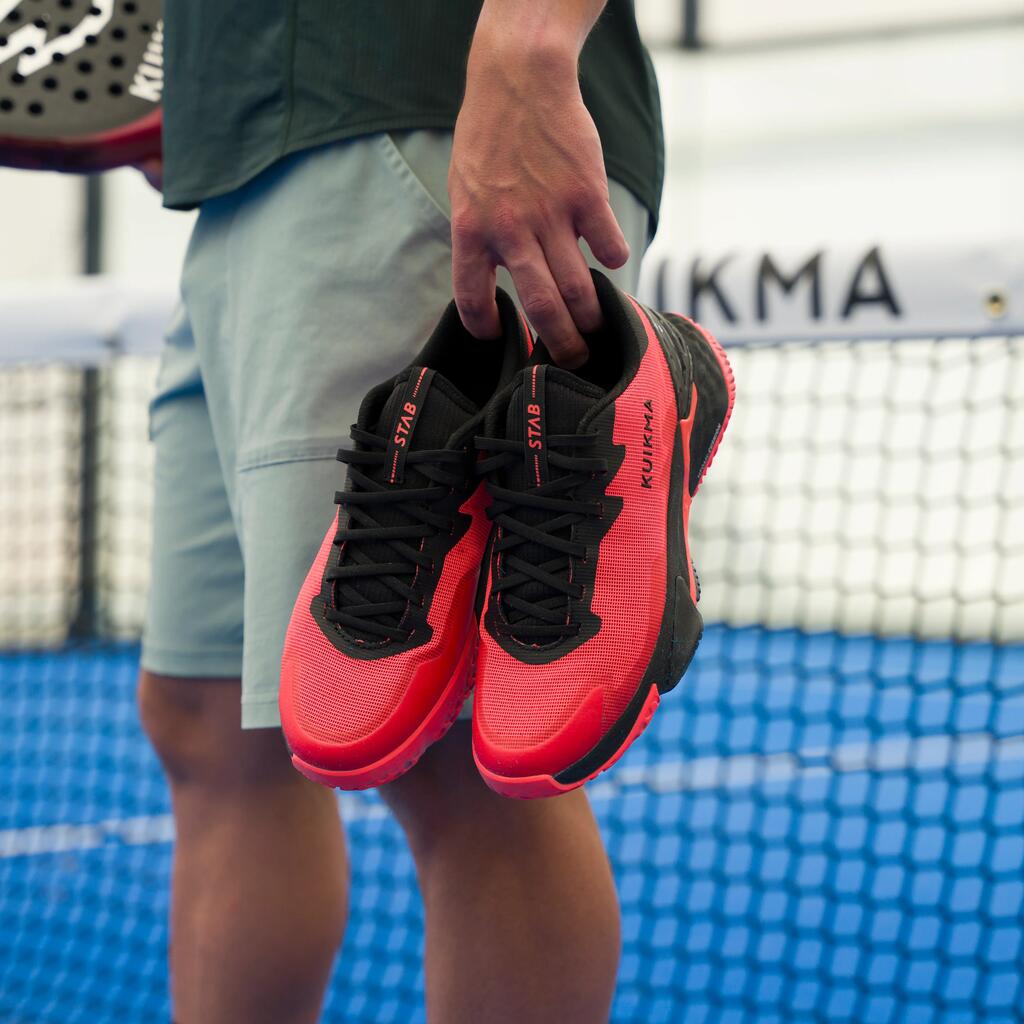 Eine Person auf einem Padel-Court hält ein paar rote Padelschuhe in der Hand; im Hintergrund ist das Netz und ein Schläger.