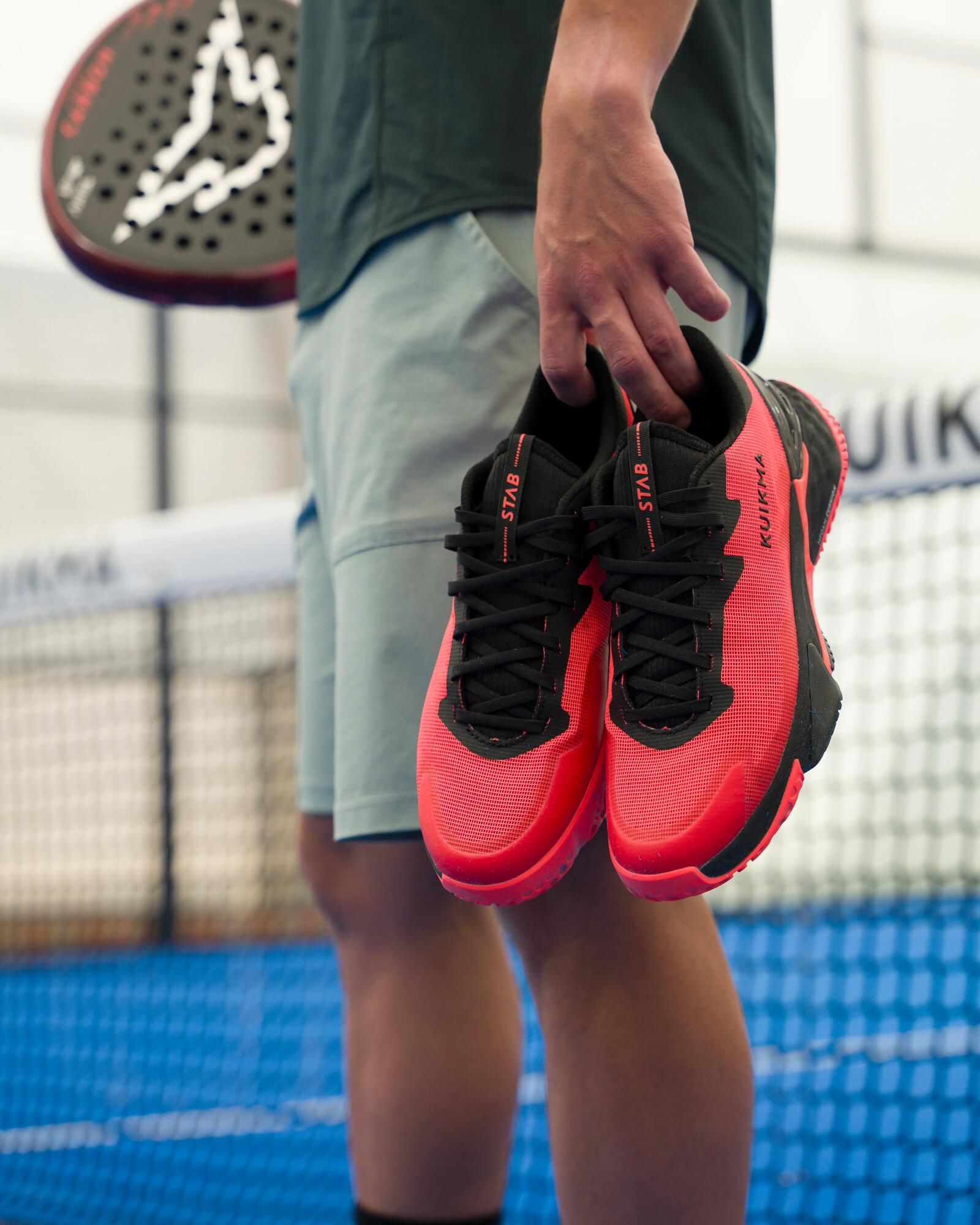 Eine Person hält ein Paar rot-schwarze Kuikma-Sportschuhe vor einem Padel-Spielfeld in der Hand.