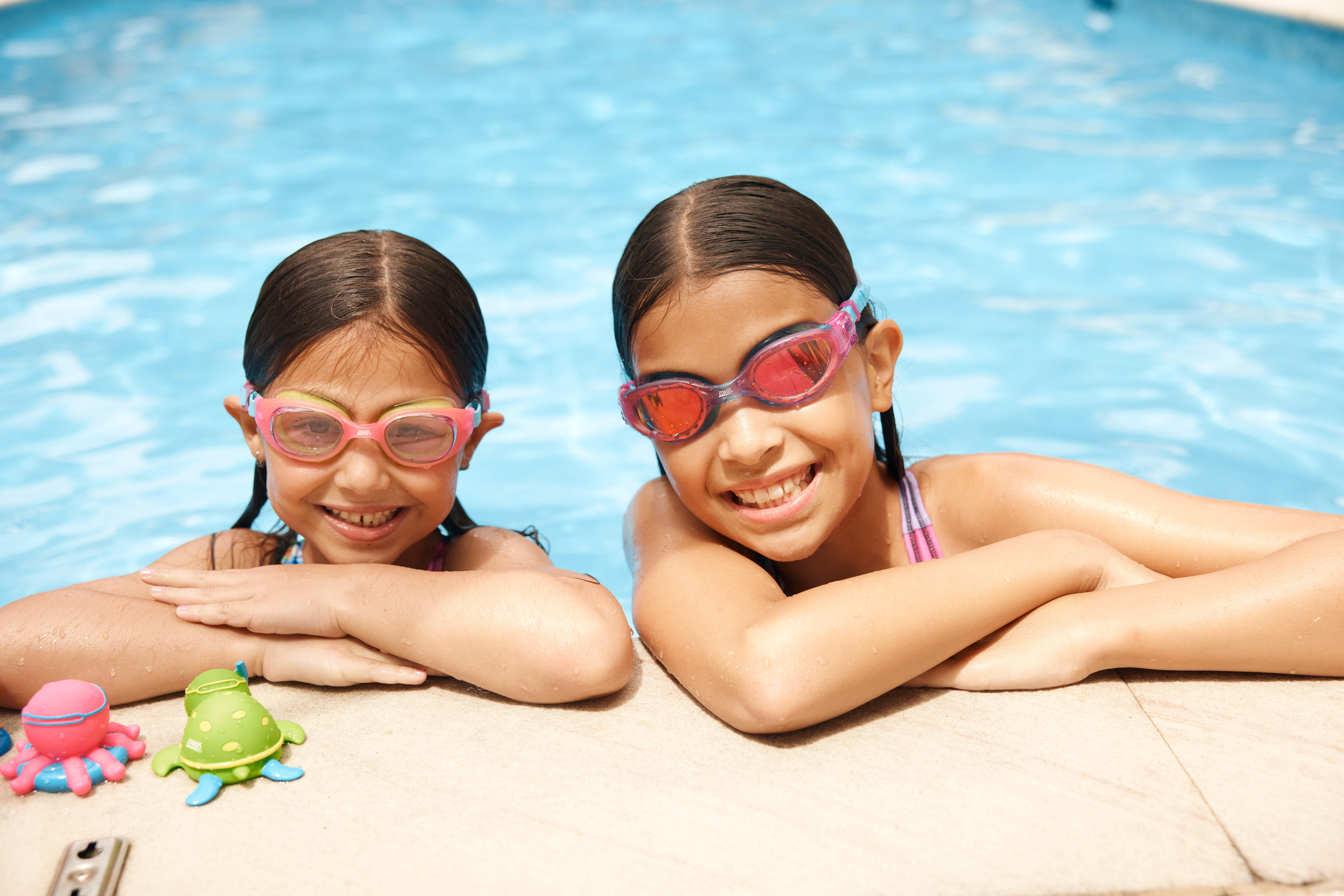 Two children wearing kids swimming goggles at the edge of a swimming pool.