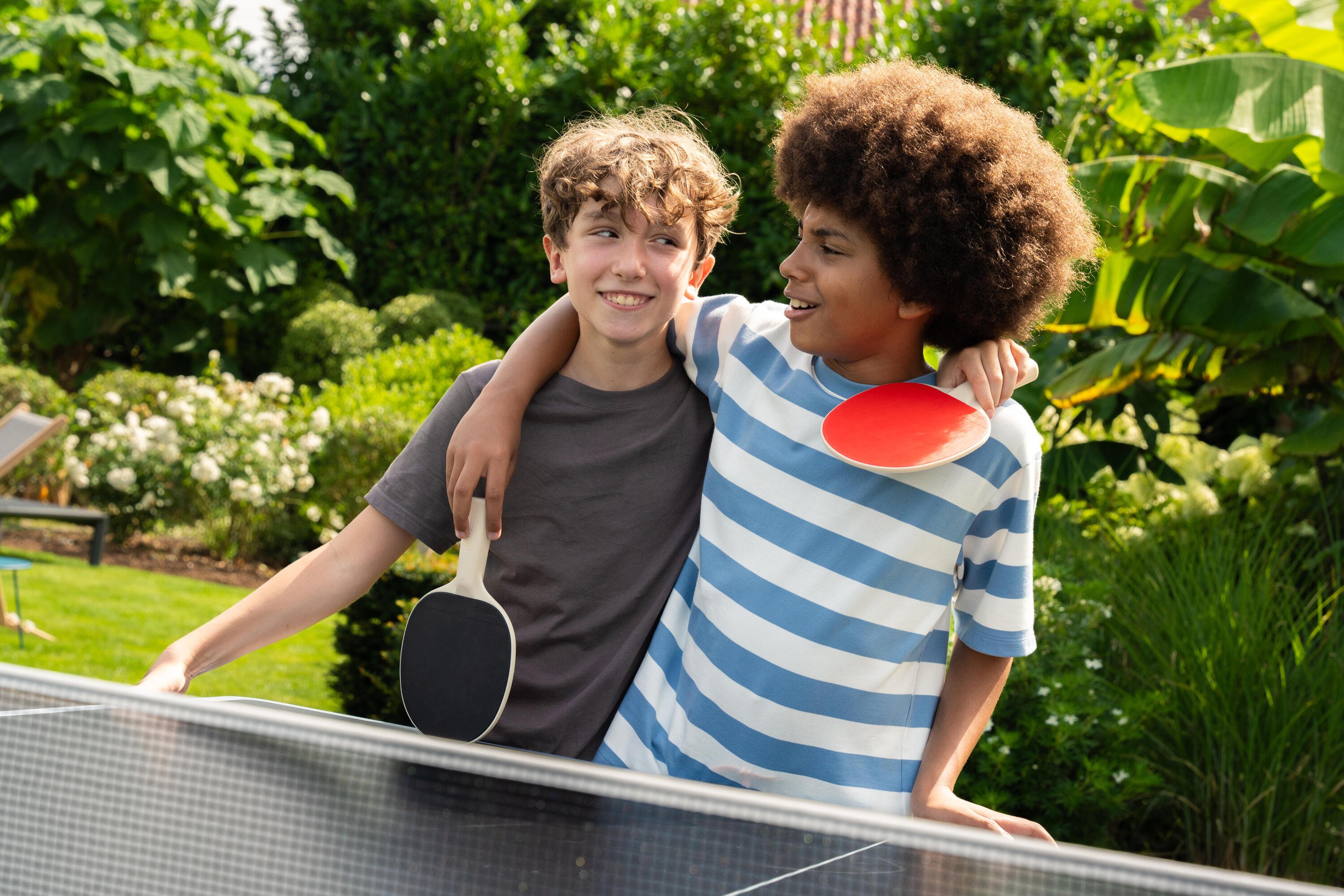 Zwei Jungen stehen lächelnd und sich umarmend mit Tischtennisschlägern an einer Tischtennisplatte im Garten.