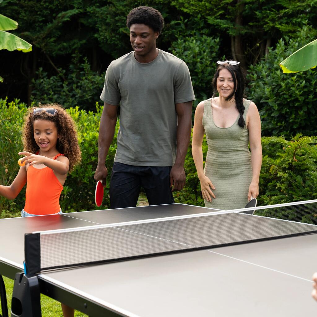 Eine Familie spielt draußen im Garten Tischtennis; ein junges Mädchen im orangenen Oberteil bereitet fröhlich ihren Aufschlag vor.