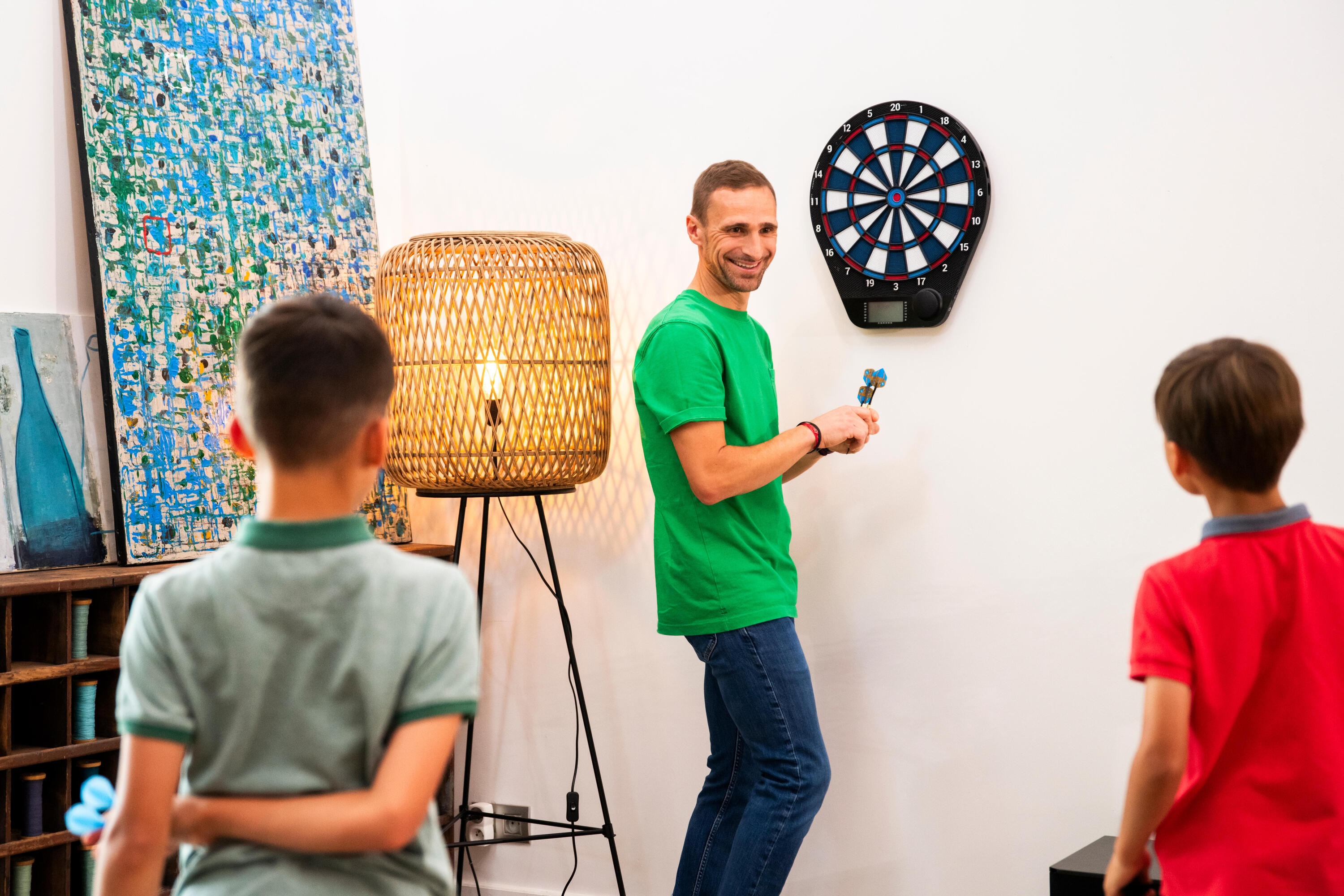 Ein Mann steht lächend vor einer elektronsichen Dartscheibe mit Darts in der Hand, während er sich zu 2 Jungs umdreht.