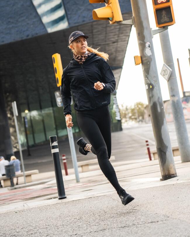 Frau beim Joggen in schwarzer Sportkleidung, gelbe Ampel, moderne Architektur im Hintergrund.
