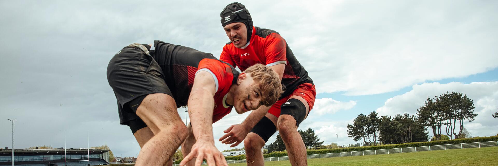 Zwei Rugbyspieler in roten Trikots kämpfen auf grüner Wiese um einen Ball. Wolkiger Himmel im Hintergrund.