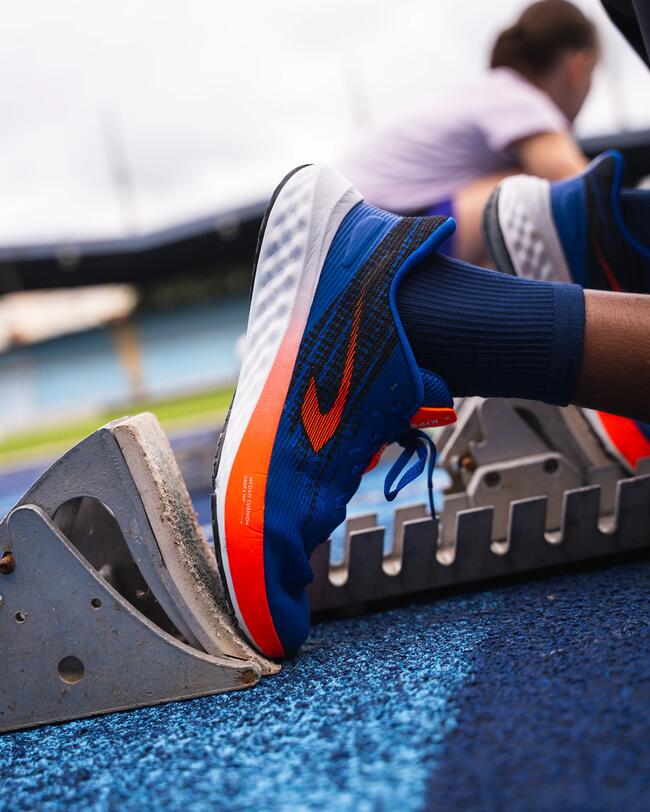 Blauer Sportschuh mit orangefarbenem Logo, steht auf Startblock vor blauem Boden, unscharfer Hintergrund.