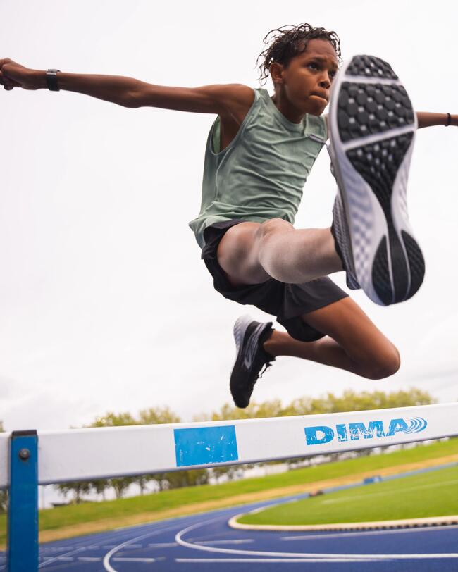 Sportlich springender Junge über Hürde, blau-grüner Sportplatz, dynamische Bewegung, helles Tageslicht, fokussierte Aktion.