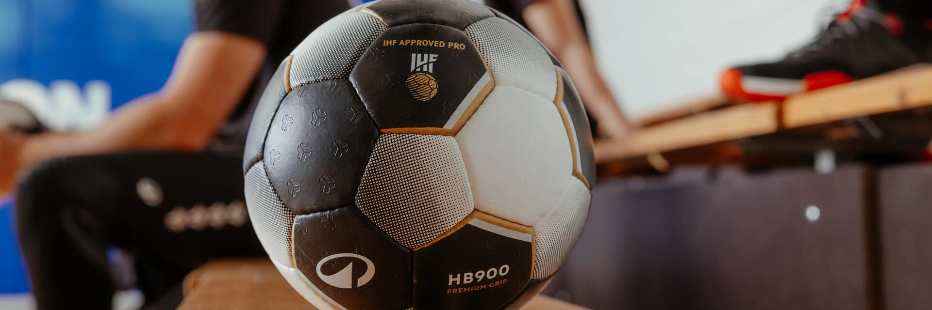 Handballspieler in schwarzer Sportkleidung, hält blau-weißen Ball, verschwommener Torwart im Hintergrund.