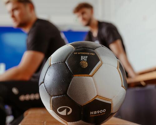Schwarz-weißer Handball auf Holzbank, unscharfe Spieler im Hintergrund, blaue Fläche.