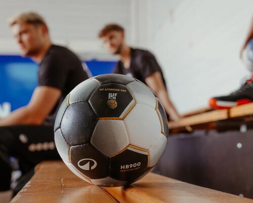 Handballspieler in schwarzer Sportkleidung, hält blau-weißen Ball, verschwommener Torwart im Hintergrund.