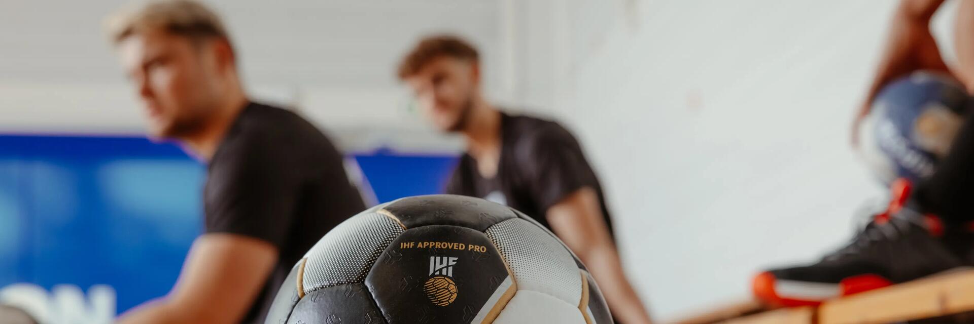 Handballspieler in schwarzer Sportkleidung, hält blau-weißen Ball, verschwommener Torwart im Hintergrund.