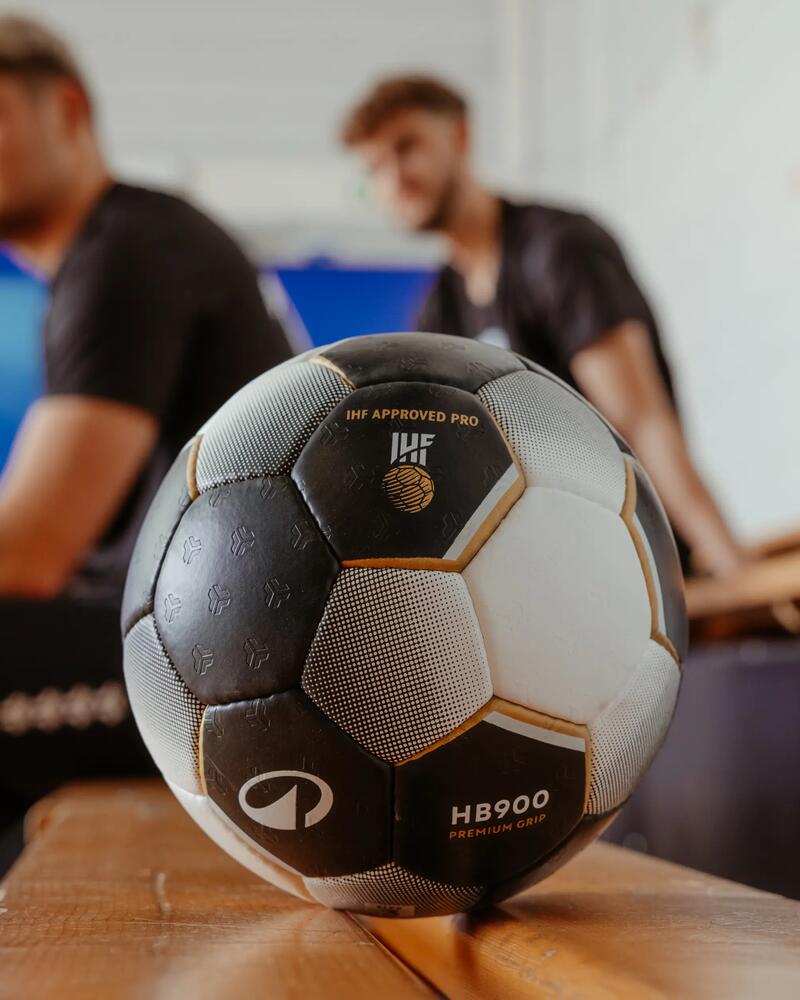 Schwarz-weißer Handball auf Holzbank, unscharfe Spieler im Hintergrund, blaue Fläche.