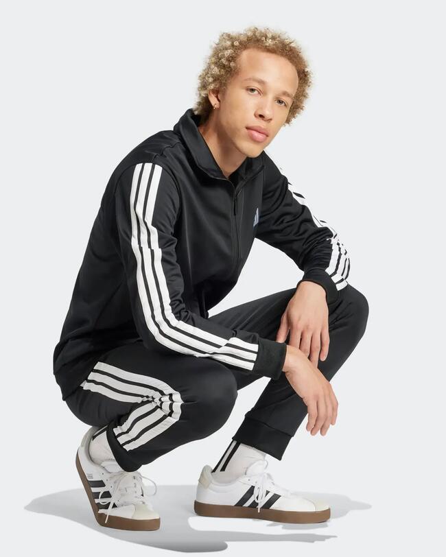 Junger Mann in einer schwarzen Adidas-Trainingsjacke und passender Hose mit weißen Streifen, hockend.