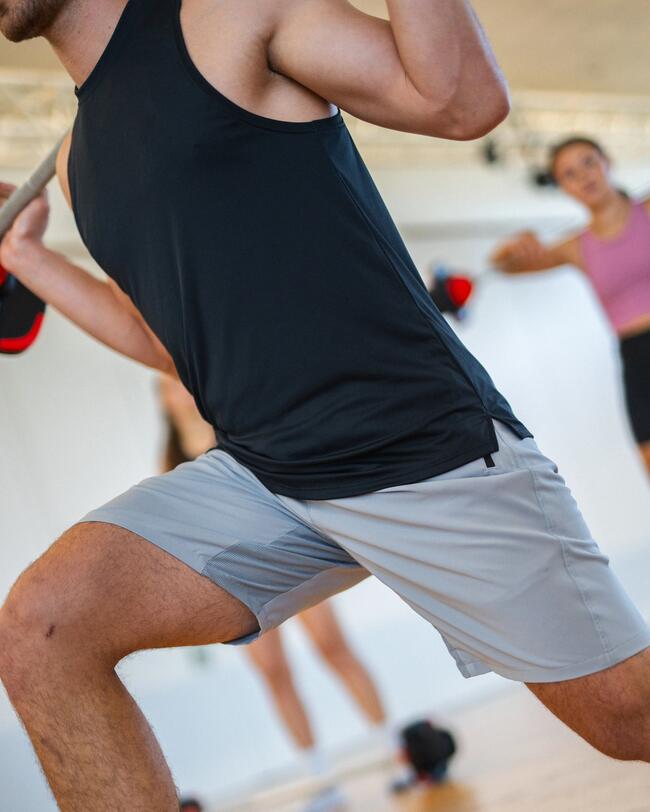 Fitness-Teilnehmer in schwarzem Tanktop und grauen Shorts hält eine Langhantel.