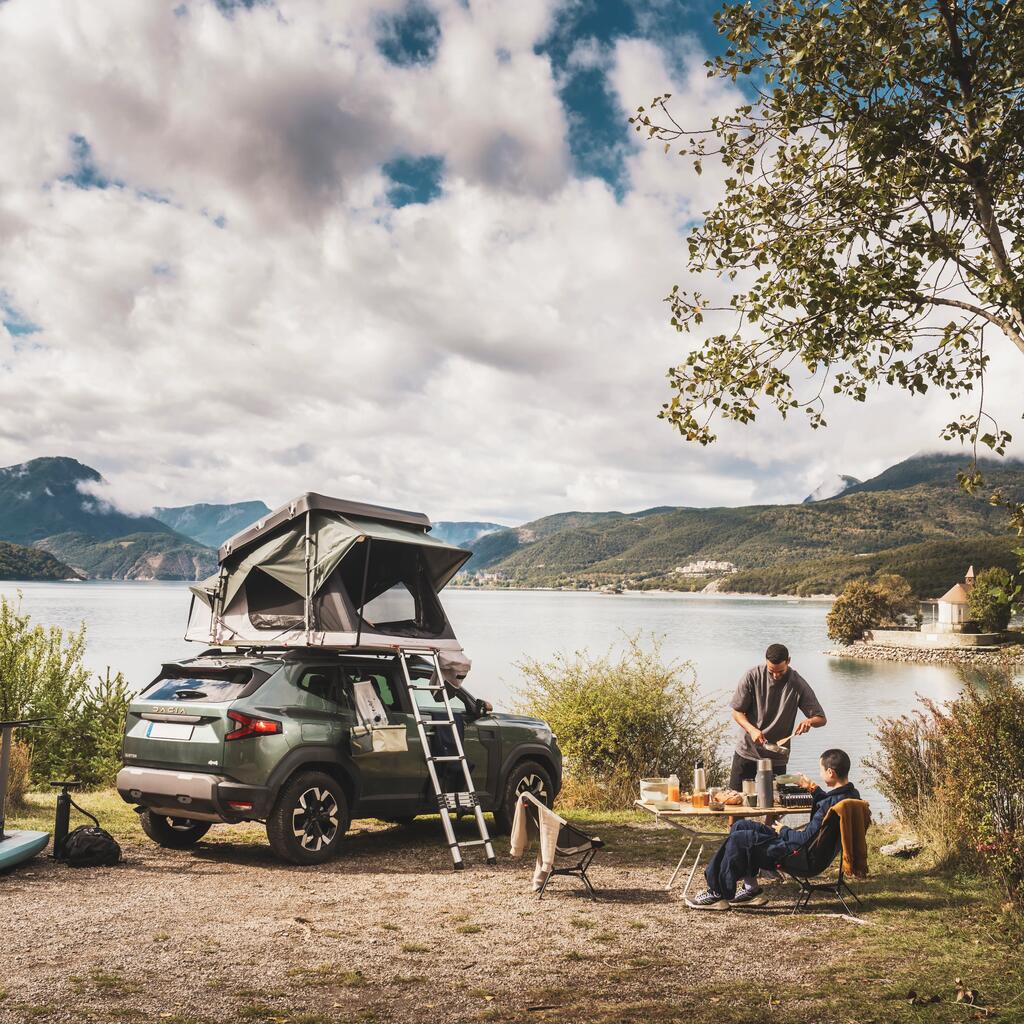 Zeleno SUV vozilo s postavljenim Quechua strešnim šotorom parkirano ob jezeru, medtem ko par pripravlja zajtrk na zložljivi mizi.
