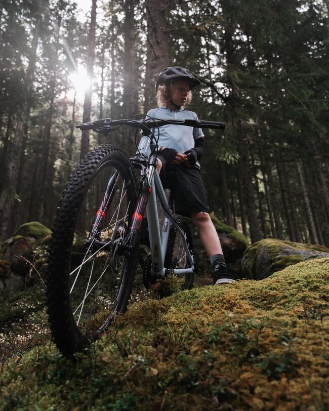 Kind mit schwarzem Helm und blauem Trikot auf grauem Mountainbike im Wald stehend