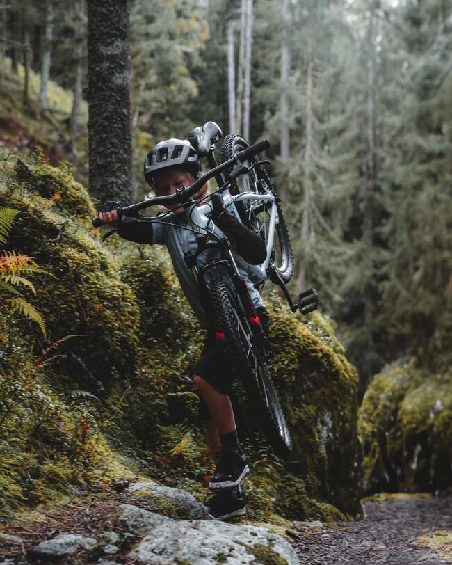 Kind mit blauem Shirt und schwarzem Helm trägt graues Fahrrad über unebenes Gelände im Wald