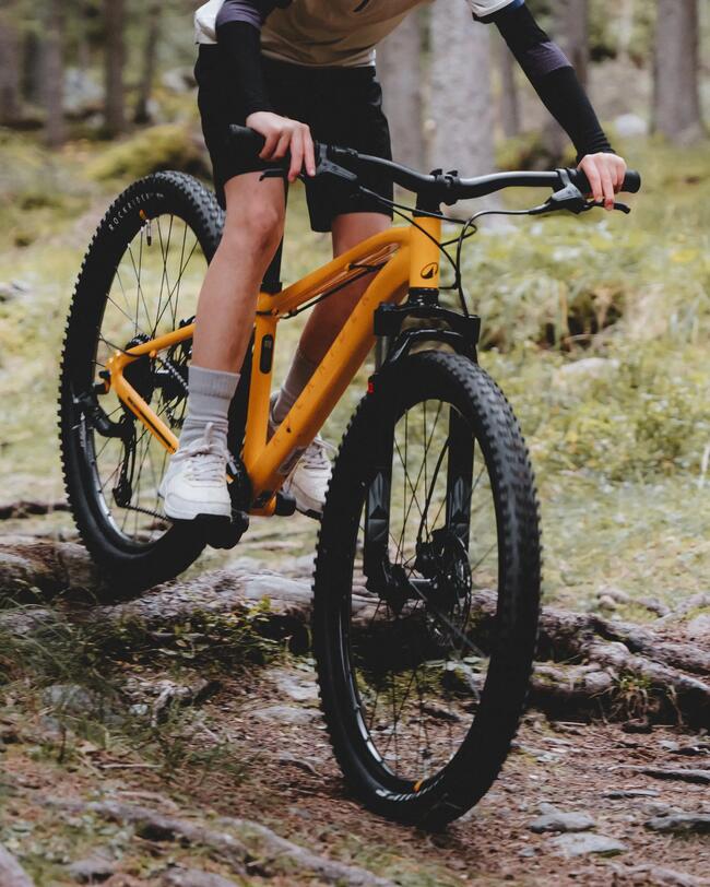 Kind fährt auf orangegelbem Mountainbike einen verwurzelten Waldweg entlang und bremst dabei