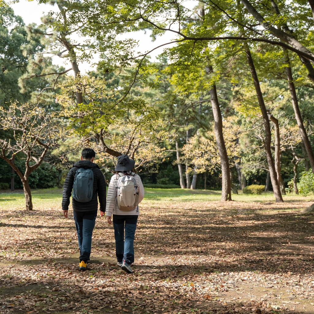 Two hikers carrying backpacks walk side by side along a leaf-covered path in a lush forest.