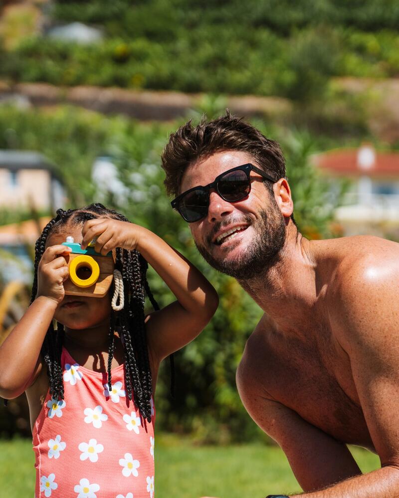 Ein lächelnder Mann mit Sonnenbrille steht neben einem kleinen Mädchen, das spielerisch durch eine gelbe Spielzeug-Kamera blickt, vor einem sonnigen Gartenhintergrund.