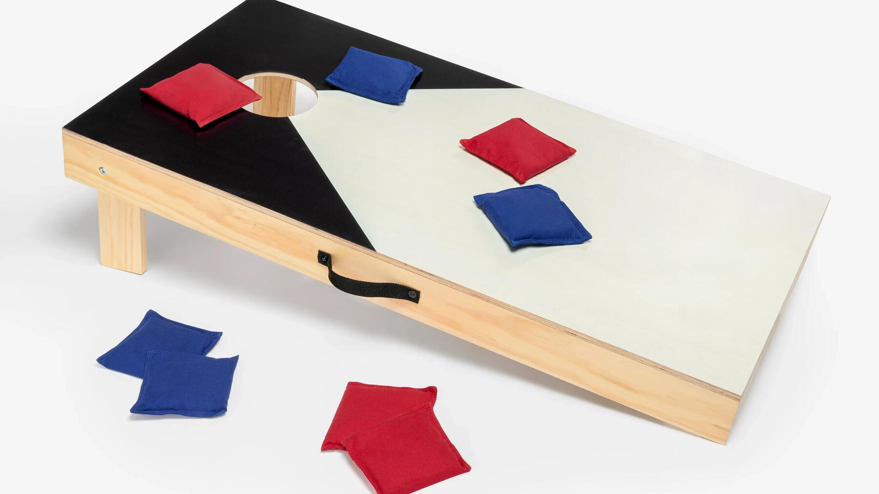 Planche inclinée en bois de cornhole mesurant 118 cm sur 60 cm avec son ouverture circulaire supérieure de 15 cm, posée sur gazon avec quatre sacs de lancer rouges et quatre sacs bleus en coton.